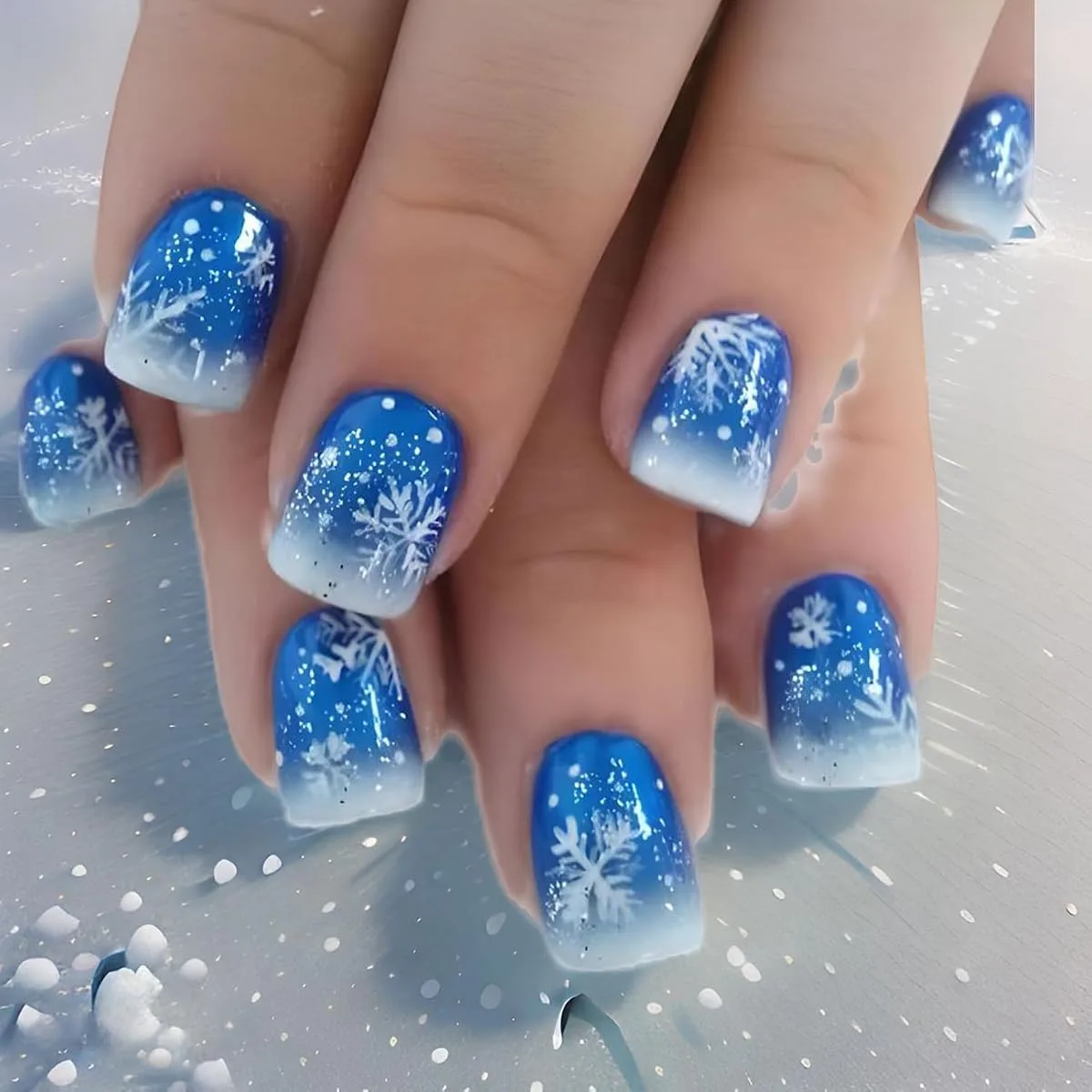 Hands with short square blue to white gradient ombre nails with white snowflakes and glitter dots on a snowy surface